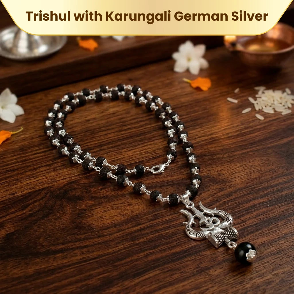 Detailed close-up of Karungali Mala showing silver-capped black ebony beads and ornate German silver Trishul pendant with OM motif at the crown — displayed on dark wood with rice grains and marigold petals