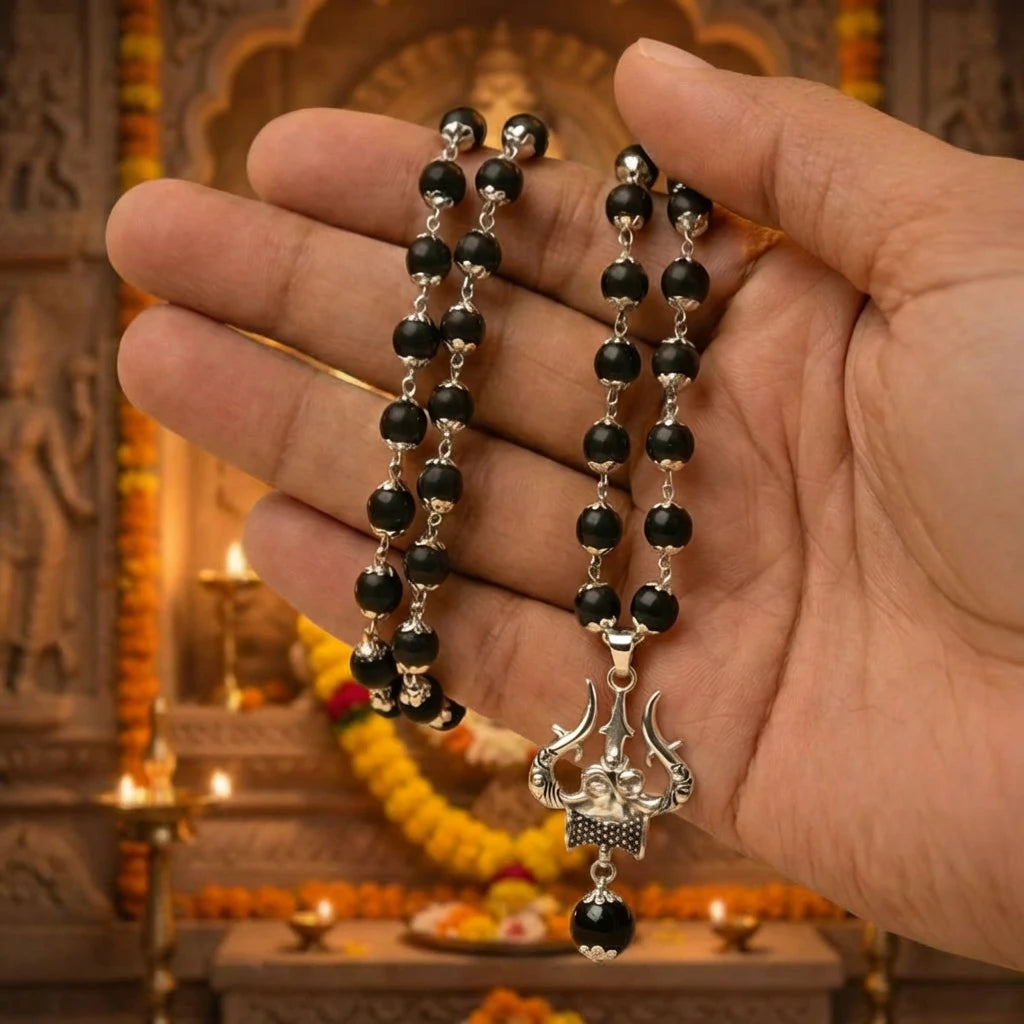 Indian male hand holding the Karungali Mala with silver Trishul pendant in front of temple altar with marigold garlands and burning brass diyas — devotional pooja context