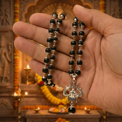 Indian male hand holding the Karungali Mala with silver Trishul pendant in front of temple altar with marigold garlands and burning brass diyas — devotional pooja context