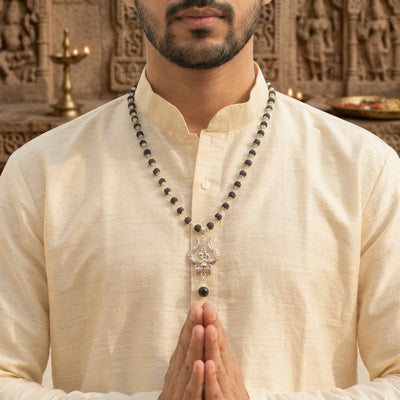 Indian man in beige cotton kurta wearing the Karungali Mala with silver Trishul pendant, hands joined in namaste in front of temple stone carving with brass diya — devotional lifestyle shot of the mala in wear